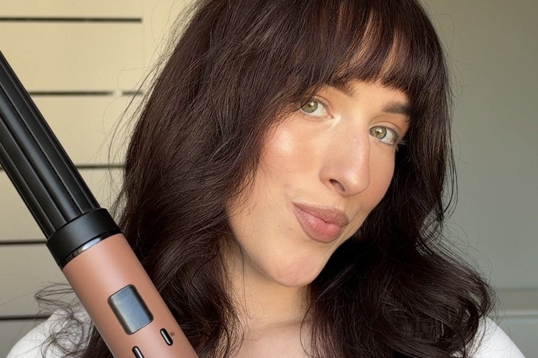 A woman with wavy hair and bangs holds the SkinVibe Nuvia AirPlasma hot air styler with a curling barrel attachment